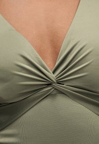 Κοντινό πλάνο ενός λεπτομέρειας ελαφρώς πράσινου υφάσματος που είναι στριμμένο και δημιουργεί μια λαιμοκοπτική γραμμή σε σχήμα V πάνω σε λεία επιδερμίδα.