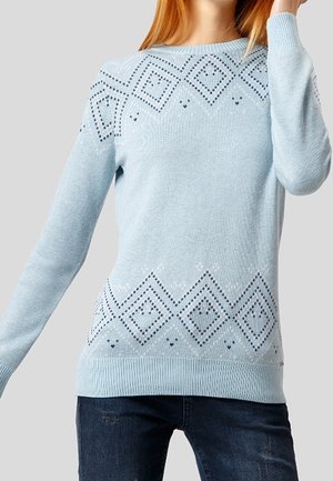 Jasnoniebieski sweter z dzianiny z granatowymi wzorami w kształcie diamentów na klatce piersiowej i u dołu, noszony z ciemnoniebieskimi dżinsami.
