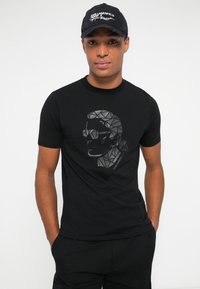 Hombre con gorra negra de Karl Lagerfeld y camiseta negra con un diseño geométrico de la cara de Karl Lagerfeld, de pie con las manos en los bolsillos.