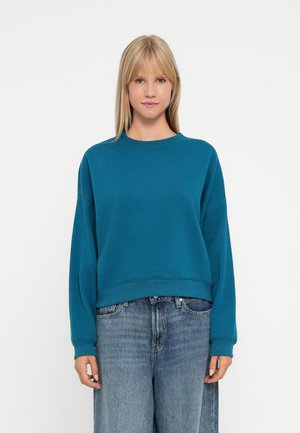 Teal genser med rund hals, senkede skuldre og ribbestrikket kant, kombinert med baggy lyseblå denimjeans.