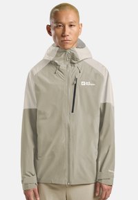 Uomo con capelli biondi corti che indossa una giacca da esterno con cappuccio di Jack Wolfskin di colore beige chiaro e bianco sporco, in piedi davanti a uno sfondo bianco semplice.
