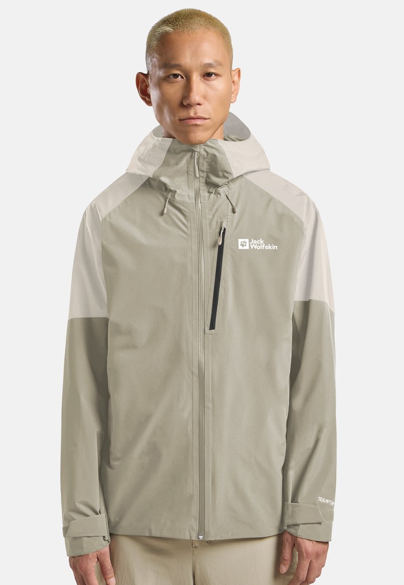 Uomo con capelli biondi corti che indossa una giacca da esterno con cappuccio di Jack Wolfskin di colore beige chiaro e bianco sporco, in piedi davanti a uno sfondo bianco semplice.
