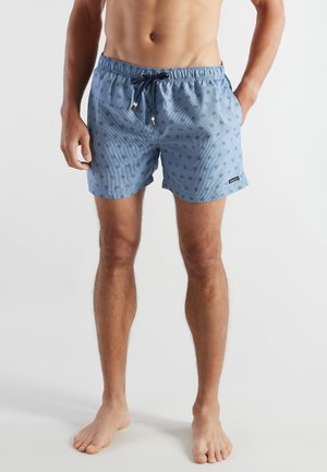 Male model wearing blue swim shorts with small dark circular patterns and a black drawstring, standing barefoot against a plain background.