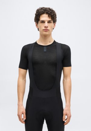 Jeune homme portant un cuissard de cyclisme noir à bretelles à manches courtes et un maillot noir ajusté en mesh, debout devant un fond clair uni.