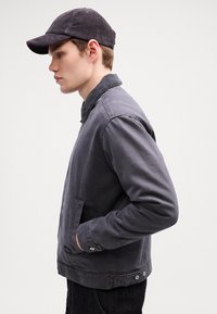 Young man in a dark gray corduroy cap and matching jacket with hands in pockets, facing left against a plain white background.