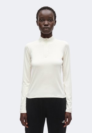 Vrouw draagt een nauwsluitende witte top met lange mouwen en een korte ritskraag, en een zwarte broek, staand tegen een effen achtergrond.