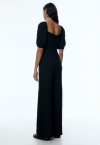 Schwarzer Jumpsuit mit Puffärmeln und quadratischem Rückenteil, aus strukturiertem Stoff und mit weitem Bein. Schlicht, monochromer Stil.