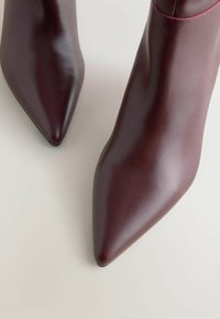 Burgundy nahkasaappaat nilkkaosuilla, joissa on terävä kärki ja sileä pinta. Ominaisuuksiin kuuluu tyylikäs muotoilu ja vähäinen ompelu yksityiskohta.