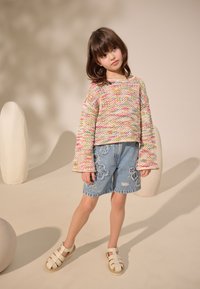 Pull en maille coloré à manches larges, shorts en denim ornés de broderies florales, et sandales blanches avec boucle. Arrière-plans simples.