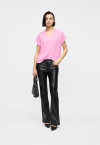 Γυναίκα που φοράει φαρδιά ροζ μπλούζα με V-λαιμόκοψη, μαύρο παντελόνι από ψεύτικο δέρμα, μαύρα μυτερά παπούτσια, κρατώντας μια μαύρη τσάντα με σχέδια.