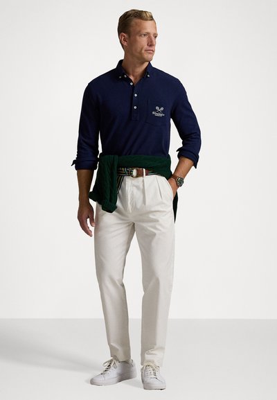 Navy long-sleeve polo shirt with a buttoned collar and emblem, cream trousers, green sweater tied at the waist, and white sneakers.