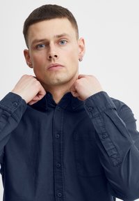 Marineblå skjorte med knapper, krage, lange ermer og brystlomme; laget av glatt stoff med en lett glans og subtil tekstur.