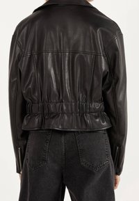 Zwarte leren jas met elastische taille en ritsmanchetten, gedragen met vervaagde zwarte denim jeans, getoond vanuit de achterkant op een neutrale achtergrond.