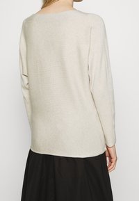 Hellbeige Strickpullover mit weitem Ausschnitt, überschnittenen Schultern und Rippstruktur, von hinten vor einem dunklen Rock gezeigt.