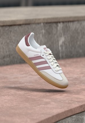 Biało-różowy but sportowy Adidas Samba z gumową podeszwą unoszący się nad kamienną powierzchnią oraz rozmytym kamiennym murem w tle.