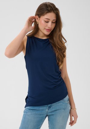 Giovane donna con capelli castani lunghi che indossa una canotta blu navy e jeans azzurri, toccandosi delicatamente i capelli e guardando verso il basso.