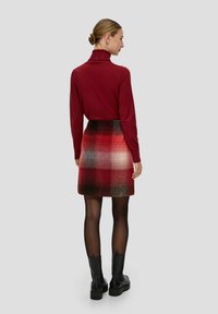 Czerwony sweter z golfem i mini spódniczka w kratę w odcieniach czerwieni, czerni i bieli. Modelka nosi czarne botki za kostkę i przezroczyste czarne rajstopy.