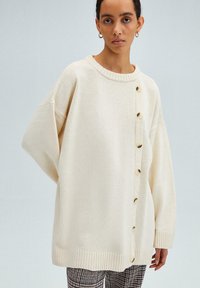 Cream-colored knit sweater with a round neck, oversized fit, ribbed cuffs, and a button-down front featuring six beige buttons.