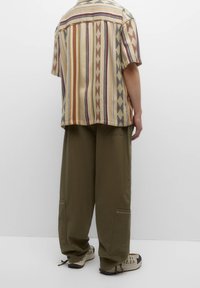 Camicia a maniche corte, multicolore, con righe verticali e motivi geometrici; abbinata a pantaloni larghi verde oliva e sneakers beige.