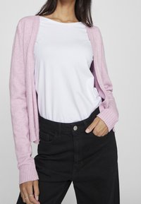 Hellrosa gestrickte Strickjacke mit gerippten Bündchen, getragen über einem weißen T-Shirt, kombiniert mit hochtaillierten schwarzen Jeans.