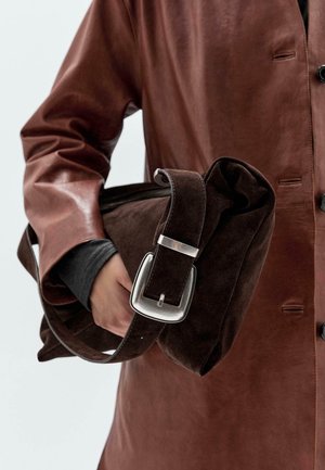 Brown suede messenger bag with a silver buckle strap, worn with a brown leather coat and dark long-sleeve top.