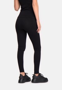 Kobieta w czarnych dopasowanych legginsach, czarnym bezrękawniku i masywnych czarnych sneakersach, stojąca z jedną ręką opartą na udzie.