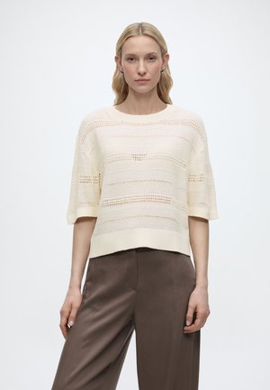 Frau mit blondem Haar, die einen cremefarbenen Strickpullover mit kurzen Ärmeln und braune, weit geschnittene Hosen trägt, vor einem weißen Hintergrund stehend.