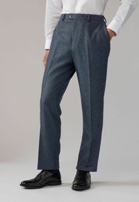Maßgeschneiderte dunkelblaue Hose mit einem subtilen gepunkteten Muster, ausgestattet mit Bundfalten, Seitentaschen und einem Knopfverschluss. Getragen mit schwarzen Schuhen.