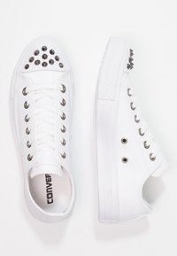 White canvas sneakers featuring silver studs on the toe, metal eyelets, and flat laces. The sole is textured and sits low to the ground.