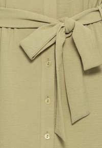 Tessuto beige testurizzato con un nodo di cintura annodata e quattro bottoni beige allineati verticalmente accanto al nodo.