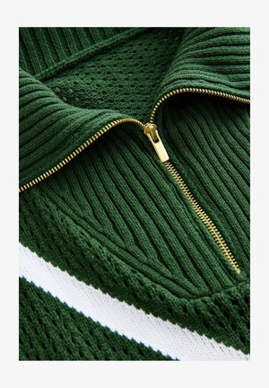 Zaļš adīts džemperis ar augstu apkakli, ar zelta rāvējslēdzēju, strukturētu ribu un baltu horizontālu joslu.