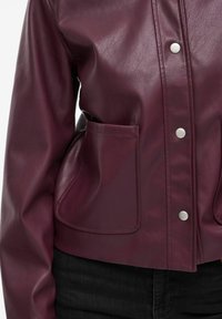 Veste en cuir bordeaux avec une texture brillante, fermeture à boutons-pression et deux poches avant. Design court et ajusté avec un minimum de détails.