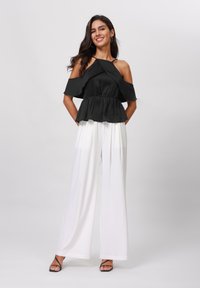 Svart off-shoulder peplum-top med samla midje, paret med flytande hvite vidbukser. Modellen står med hendene i lommene.