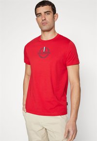 Rød bomulds T-shirt med et cirkulært marineblåt logo med laurbærdesign og to lodrette striber. Korte ærmer og rund halsudskæring.