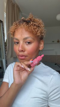 Flacon compte-gouttes rose en main, avec un bouchon texturé. La personne a des cheveux bouclés et du vernis à ongles multicolore, portant une chemise blanche.
