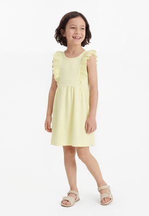 Smilende ung pige står iført en lysegul kjole med flæser på ærmerne og beige sandaler mod en ensfarvet hvid baggrund.