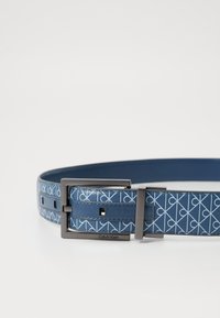 Modrý kožený opasok so svetlými geometrickými vzormi, obdĺžniková tmavá kovová pracka a rytie "Calvin Klein" na ráme pracky.