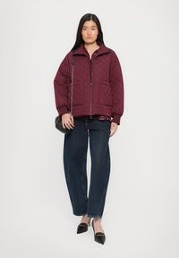 Burgundy quilted jacket with zip closure and two front pockets, paired with dark denim wide-leg pants and black pointed-toe heels.