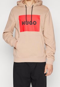 Sudadera beige con un gráfico rectangular rojo que muestra "HUGO" en una fuente negra en negrita. Cuenta con un bolsillo tipo canguro y capucha ajustable con cordón.