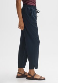 Marineblauwe, lichte broek met een elastische tailleband en trekkoord. Bevat zijzakken en een taps toelopend been met splitzoom. Gecombineerd met een paar bruine sandalen.