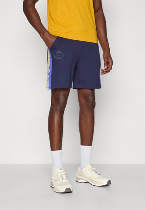 Homme portant un short Kappa bleu marine avec logo et bandes jaunes sur les côtés, chemise jaune, chaussettes blanches et baskets couleur crème, debout.
