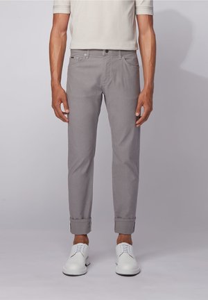 Man draagt lichtgrijze slim fit broek met opgerolde boorden en witte veterschoenen, staand tegen een effen grijze achtergrond.
