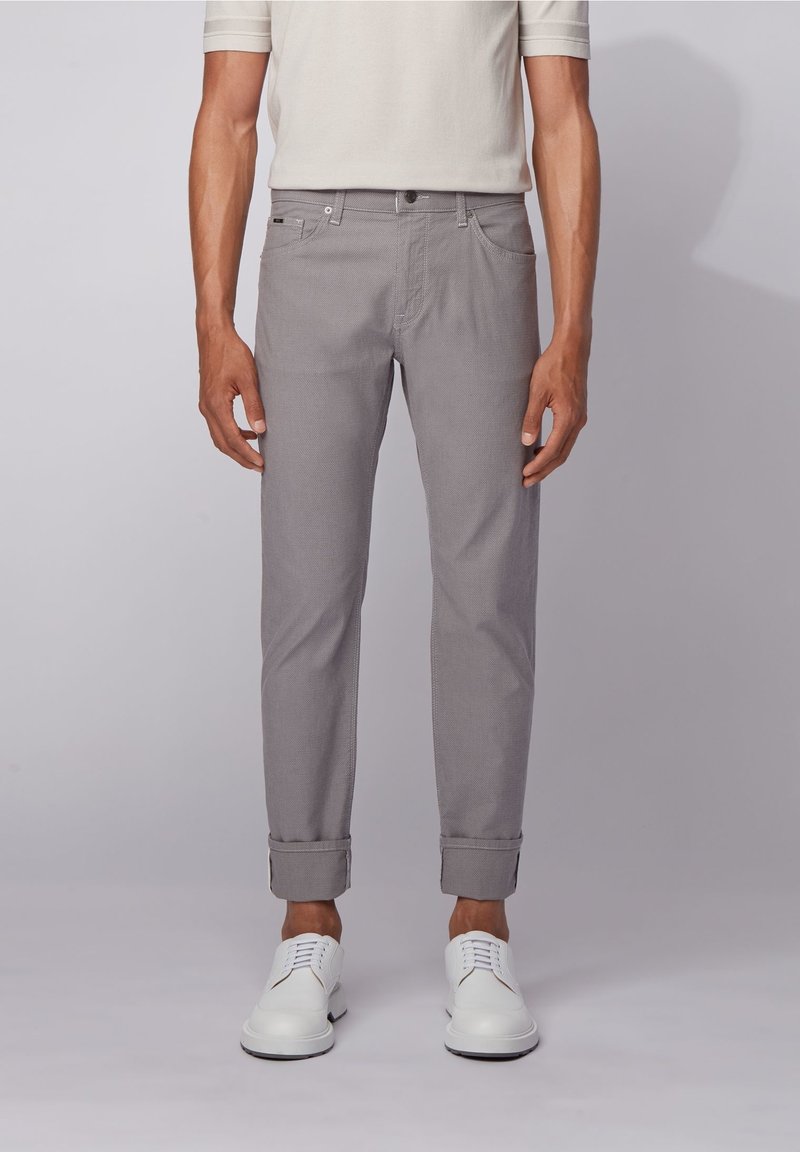 Homme portant un pantalon slim gris clair avec les revers roulés et des chaussures blanches à lacets, debout devant un fond gris uni.
