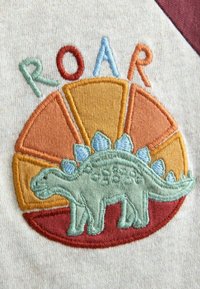 Tikkimise patch, millel on roheline dinosaurusekujuline pilt okastega, oranži, punase ja kollase tooni ringikujulise tausta ees, ning sõna "ROAR" kohal.