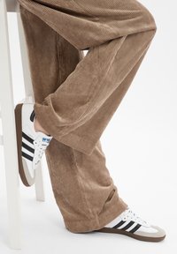 Baskets blanches et grises avec des rayures noires, semelle en caoutchouc texturé, assorties à un pantalon en velours côtelé marron à côtes verticales.