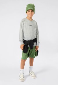 Barn som har på seg en grå langermet skjorte med en logo, grønne shorts, hvite sokker og joggesko. En svart hettegenser er knyttet rundt livet.