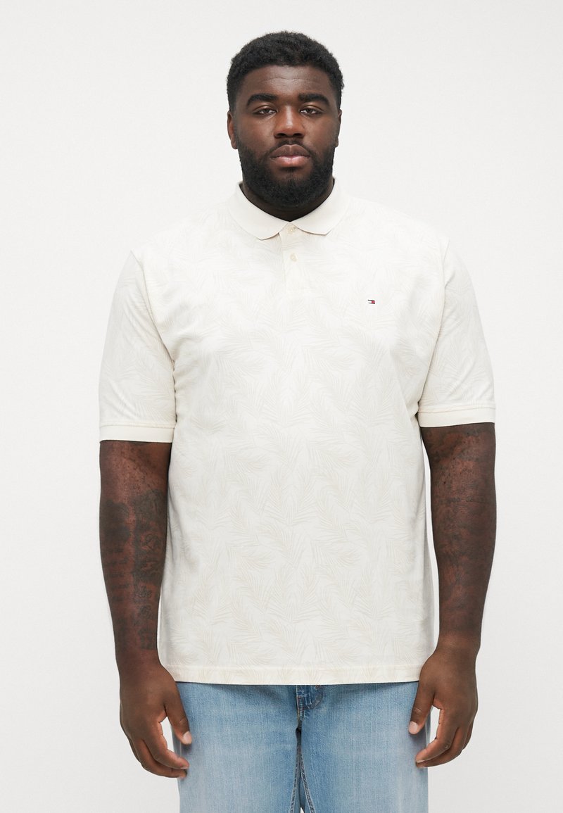 Polo a maniche corte crema con un sottile motivo di foglie, colletto abbottonato e piccolo logo sul petto. Indossato con jeans di denim blu.