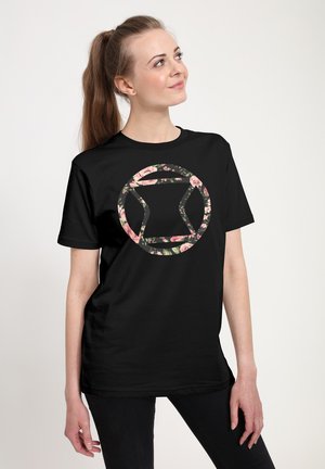 Ung kvinde med hestehale iført en sort t-shirt med et blomster-mønstrede cirkulært geometrisk design, stående foran en ensfarvet lys baggrund.