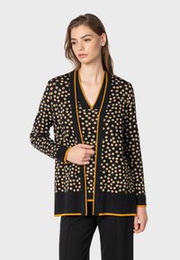 Svart kardiagan med beiga prickar och orange detaljer. Har V-ringning och långa ärmar, tillverkad av texturerat stickat material.