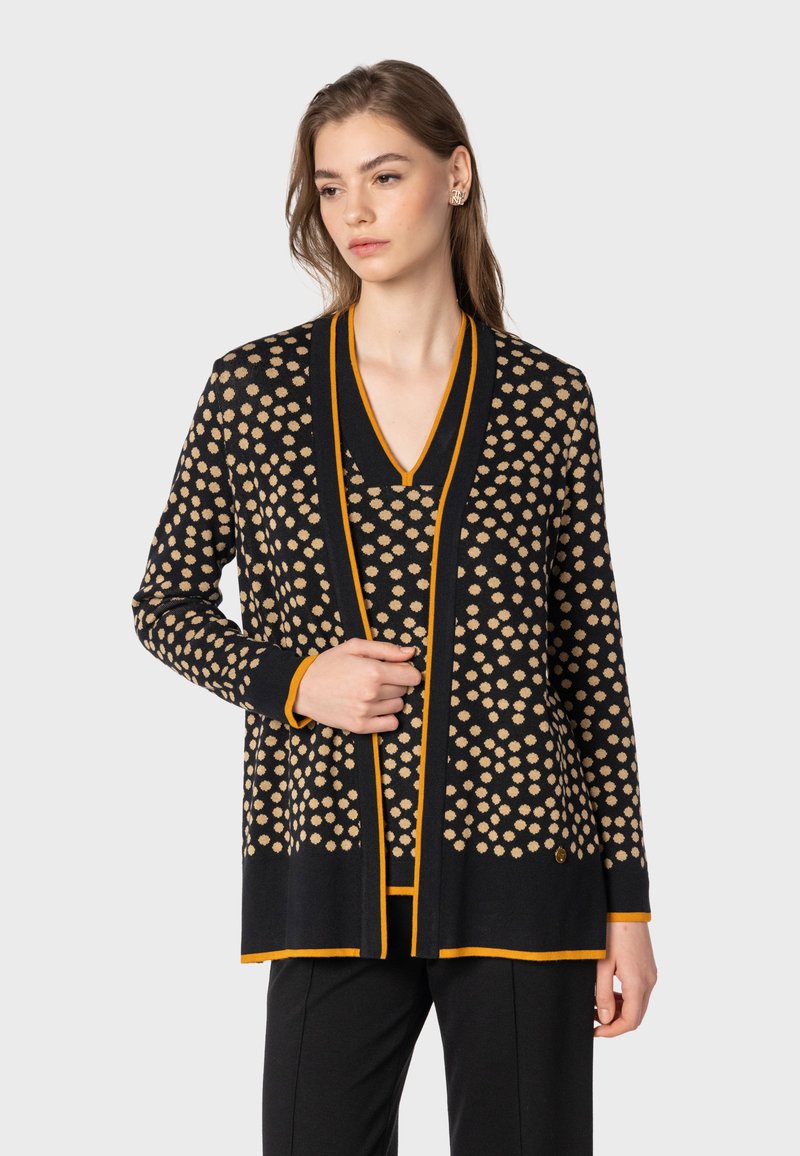 Svart kardiagan med beiga prickar och orange detaljer. Har V-ringning och långa ärmar, tillverkad av texturerat stickat material.